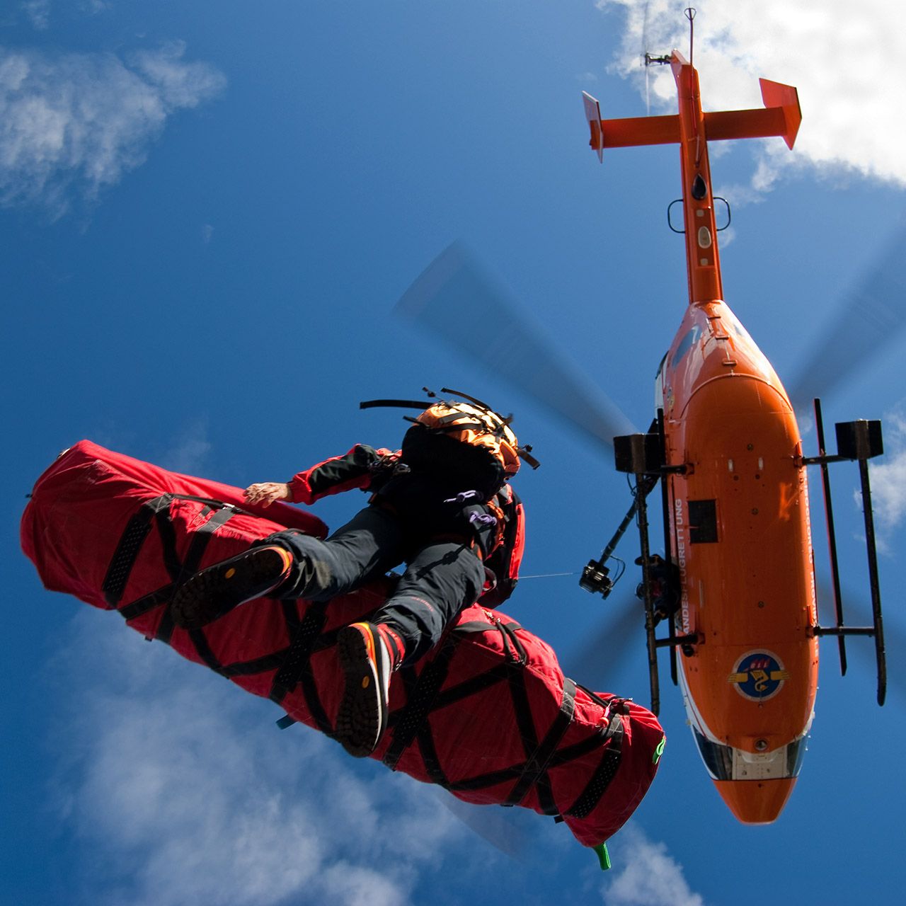 A low-angle shot of a rescue worker being airlifted by an orange helicopter while riding on a red rescue basket against a blue sky.