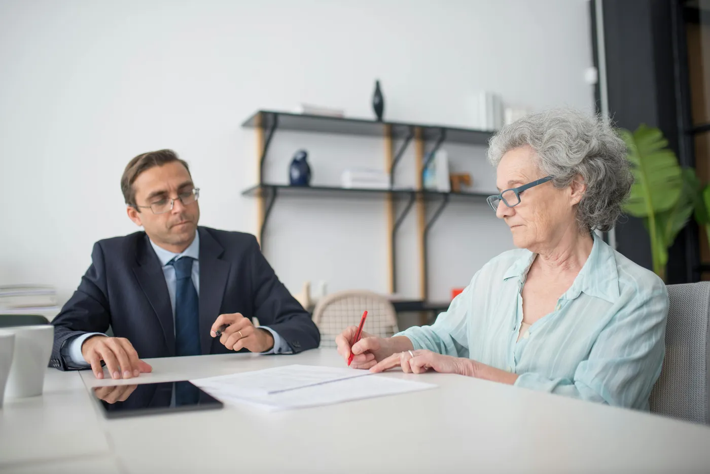 An older woman signing documents at a desk across from a suited professional in a modern office