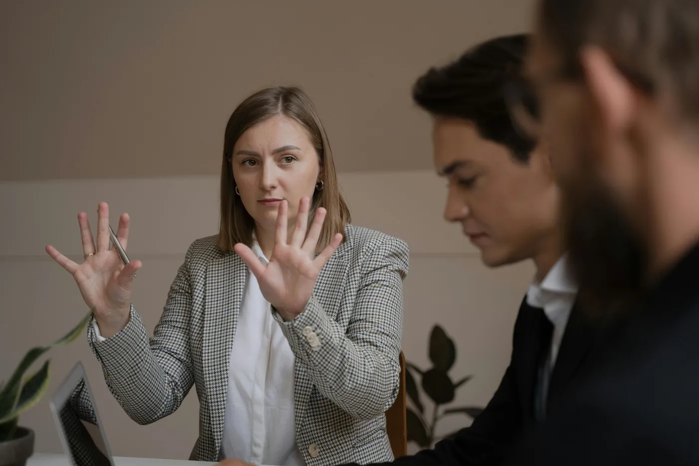 A young woman in a checkered blazer gestures with both hands as she speaks to two men in an office setting.