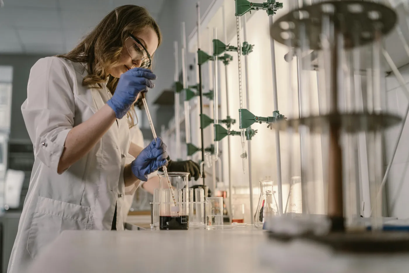 A scientist in a white lab coat and blue gloves pipetting liquid into beakers and test tubes at a laboratory bench