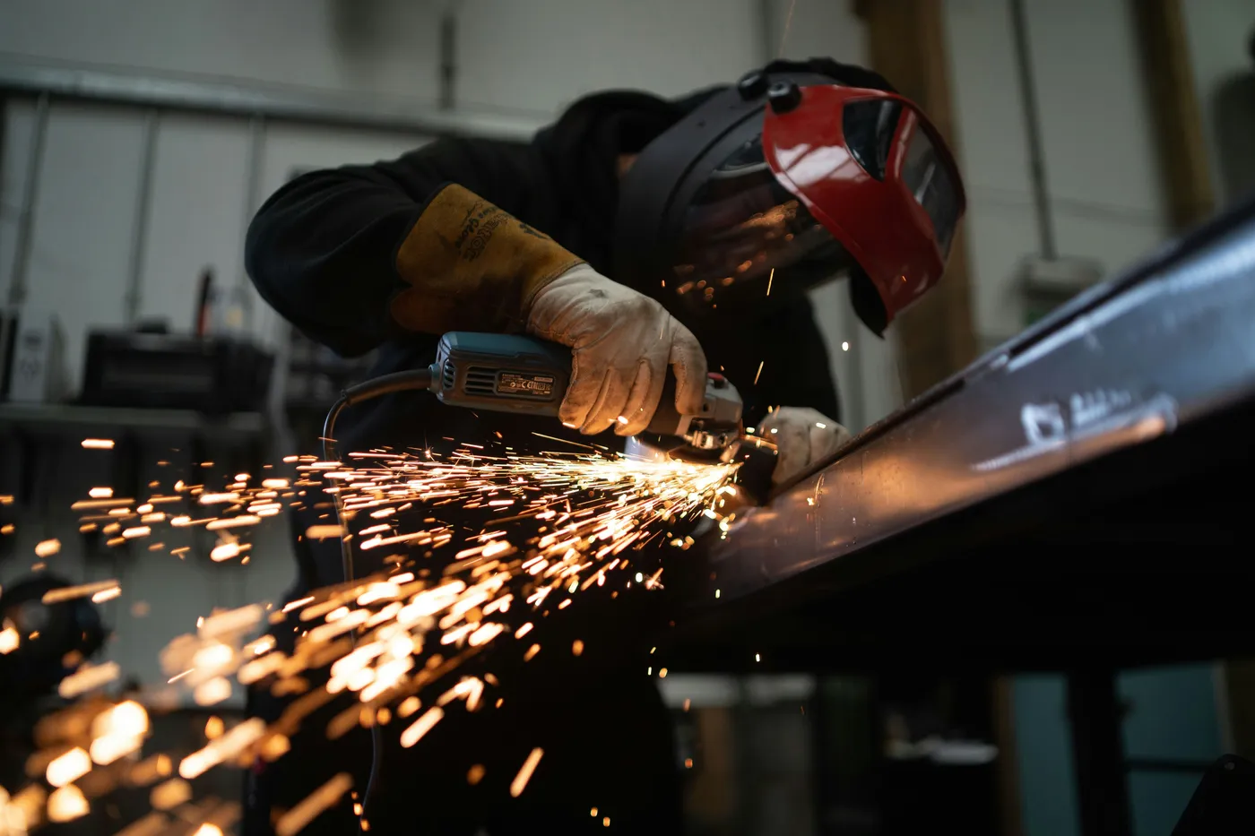 A worker in a welding mask and protective gloves grinding metal, sending a shower of sparks across a workshop floor