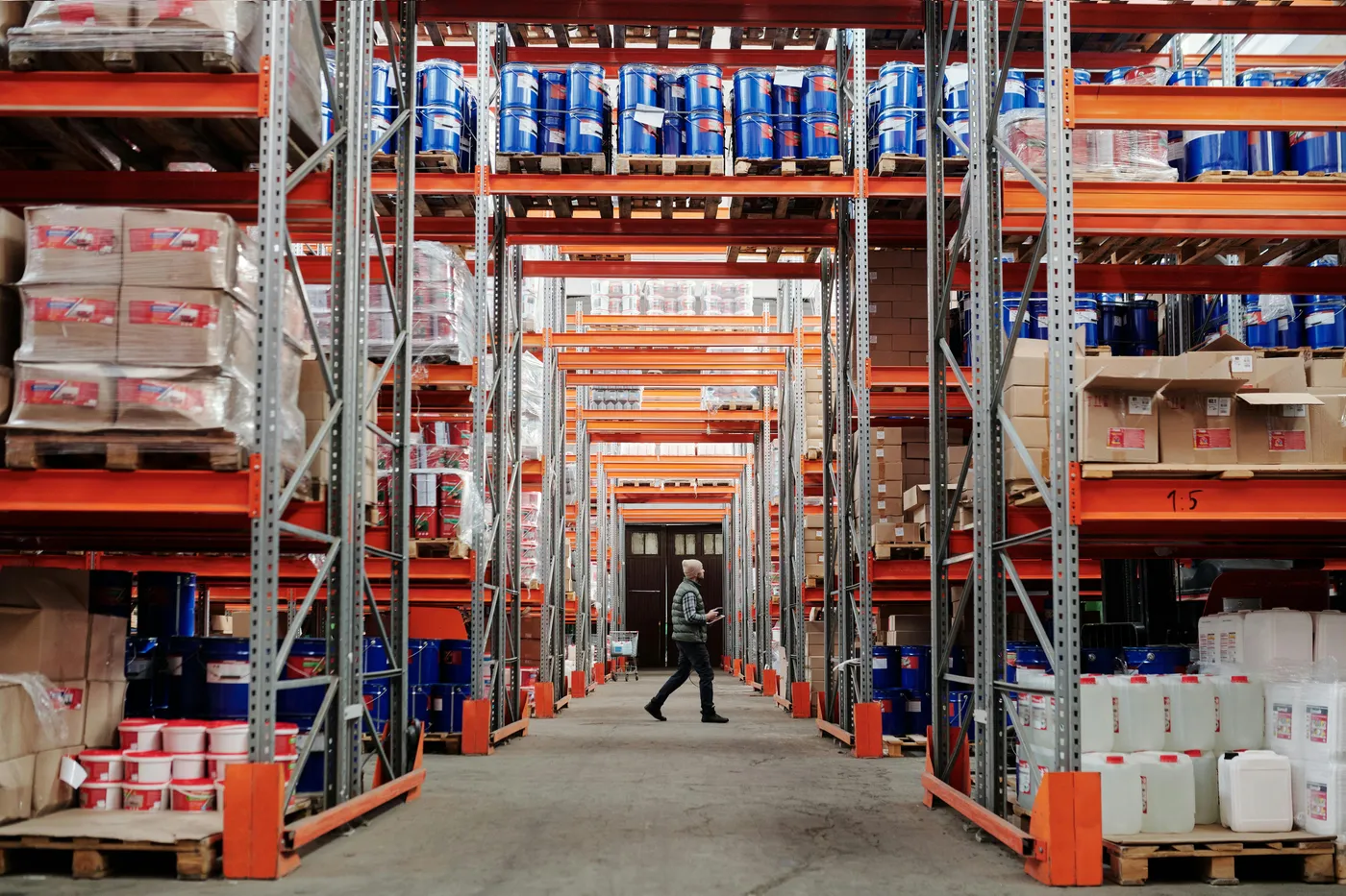A worker walking through a tall industrial warehouse aisle lined with shelving stacked with drums, boxes and containers